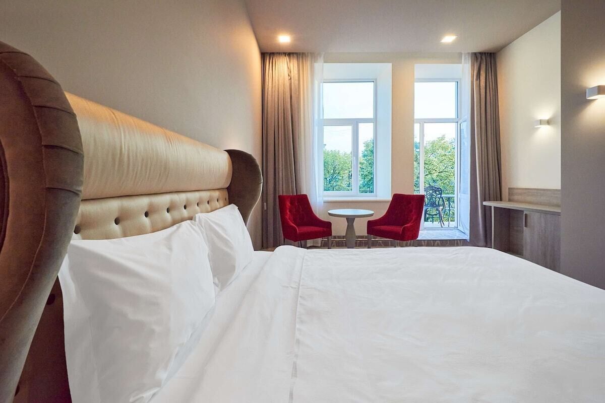 A hotel room with a large bed, a red chair, a small table, and a window with curtains.