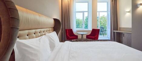 A hotel room with a large bed, a red chair, a small table, and a window with curtains.