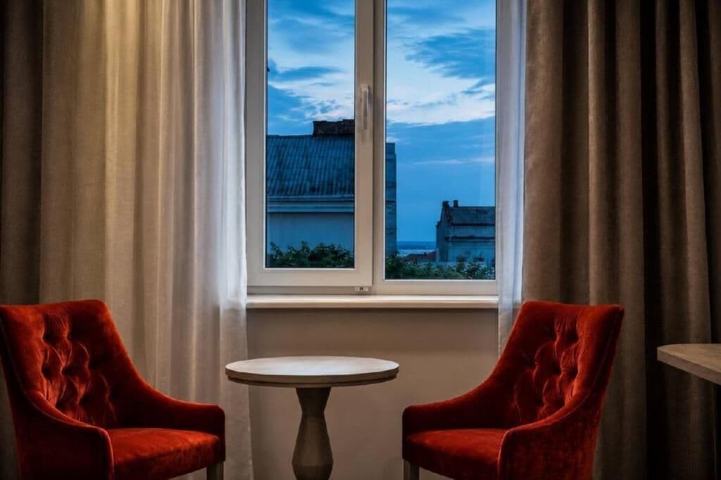 Una habitación con dos sillones de terciopelo rojo, una mesa redonda pequeña y un ventanal con vistas a edificios.