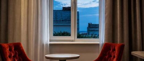 Una habitación con dos sillones de terciopelo rojo, una mesa redonda pequeña y un ventanal con vistas a edificios.