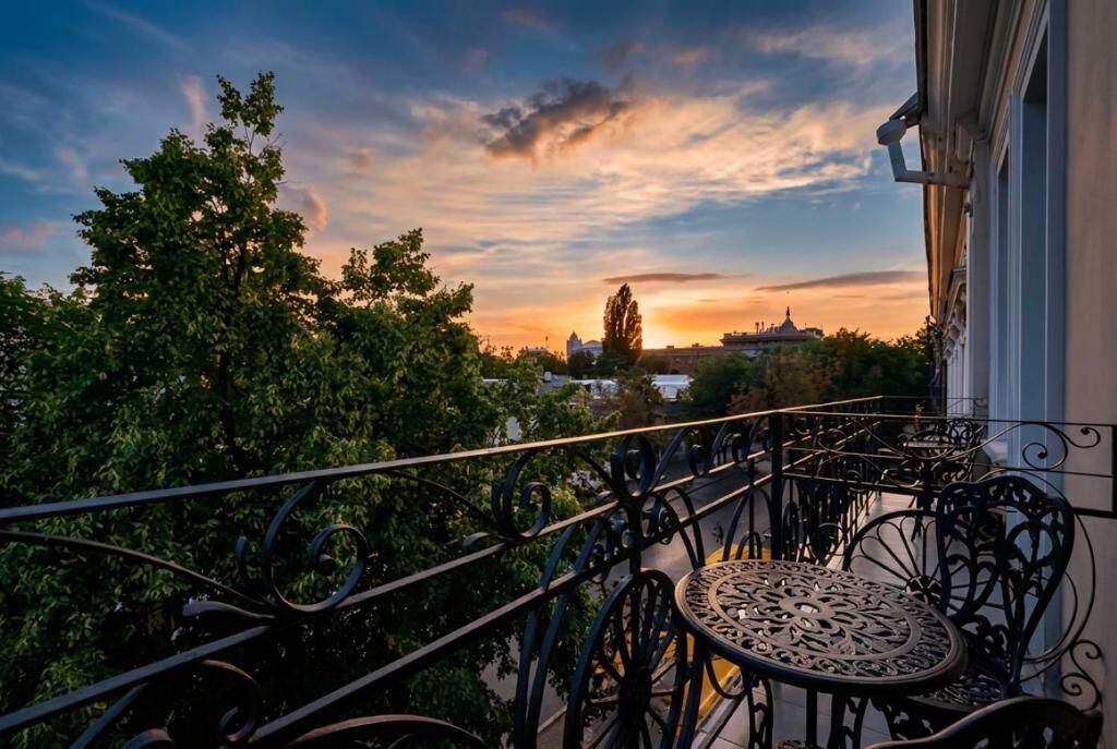 Un balcon doté de balustrades en fer forgé et d’une table, offrant une vue sur un paysage urbain avec des arbres et des bâtiments, au moment du coucher du soleil.