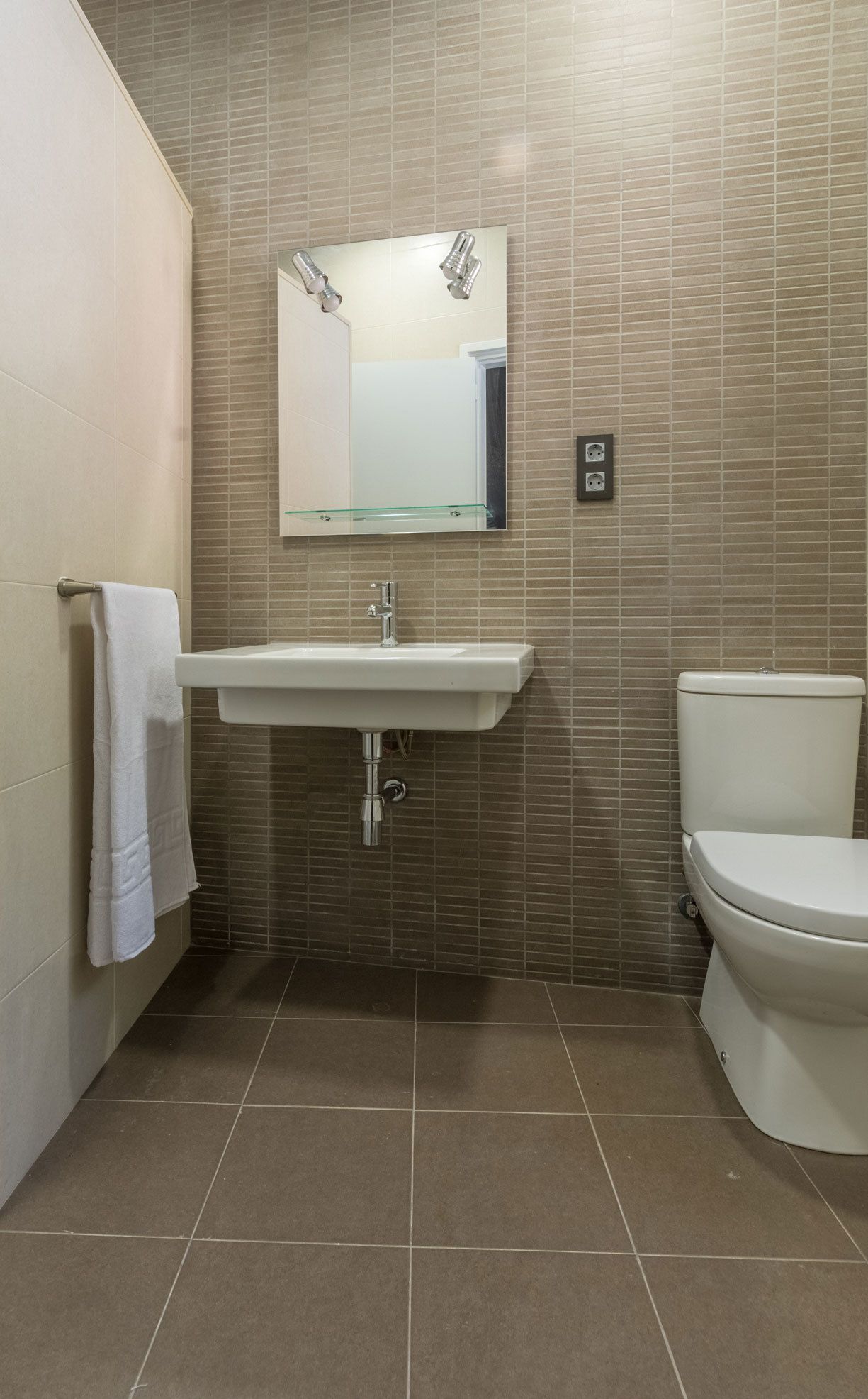 Un baño con lavamanos blanco, espejo y váter, paredes con azulejos y una toalla colgada en la pared.