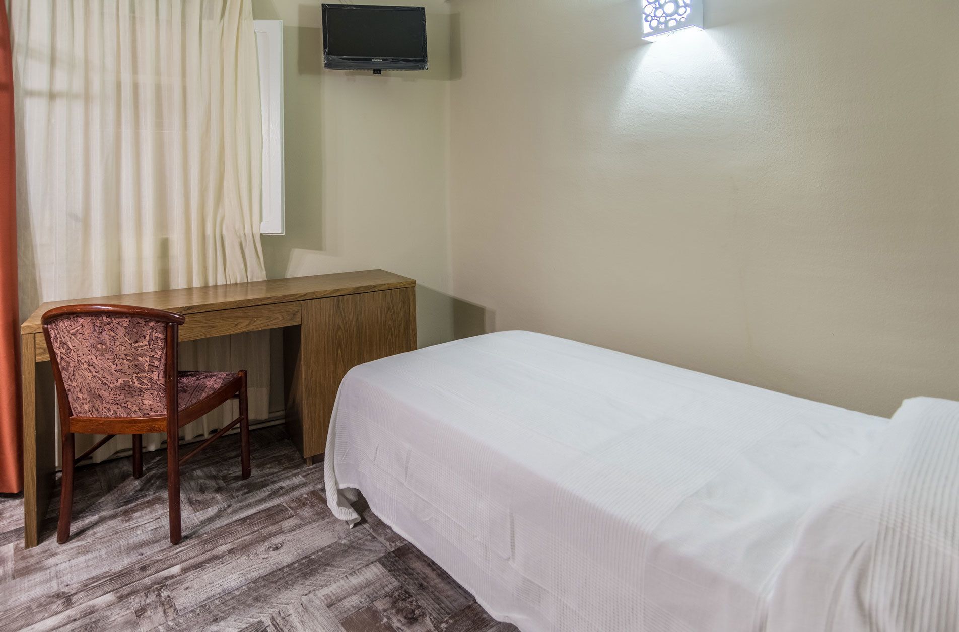 A room with a wooden desk, a single chair, a bed with a white bedspread, a television mounted on the wall, and a window with a decorative screen.