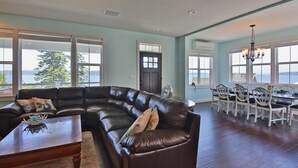 A spacious living room with a dark leather sectional sofa, a wooden coffee table, and a dining area with white chairs and a chandelier. - The Lookout at Cranes Landing (Greenbank)