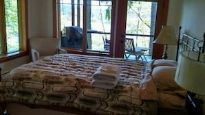 A bedroom with a large bed, a chair, a table, and a window with a view of trees. - Goss Lake Chalet (Langley)
