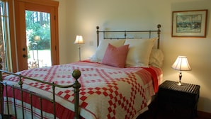 A bed with a checkered quilt, two bedside lamps, a framed picture on the wall, and a door leading to a garden. - Goss Lake Chalet (Langley)