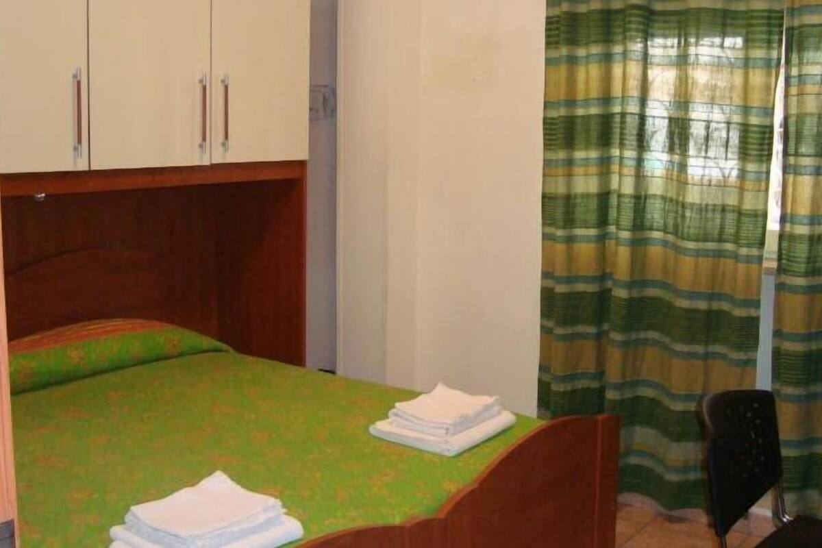 A small, single-room hotel room with a bed, a desk, a chair, and a wardrobe.