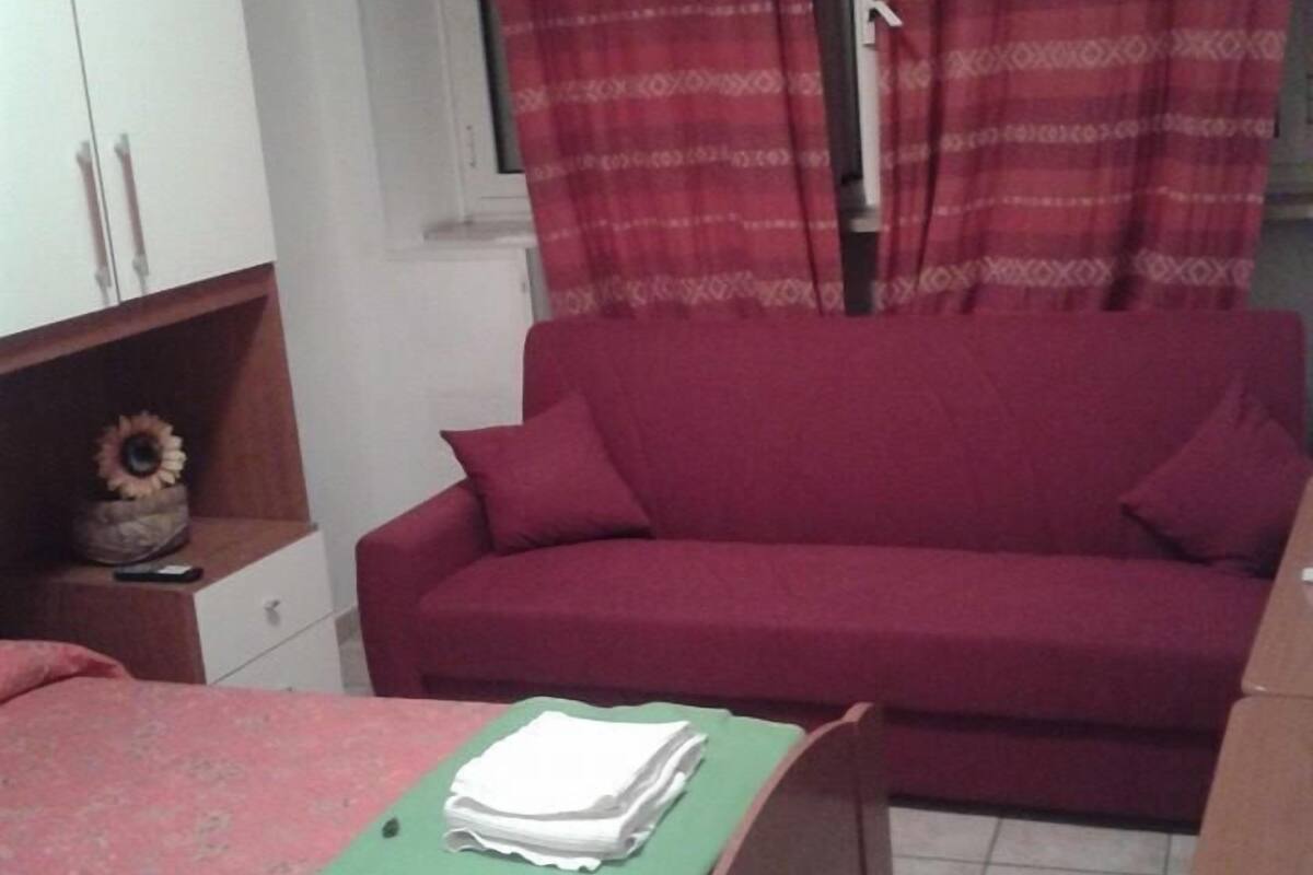 A bedroom with a bed, a red sofa, a green table with folded towels, a wooden cabinet, and a window with red curtains.