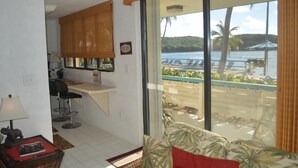 A living room with a sofa, a small table with a lamp, and a kitchen area with a counter and chairs. - 1612 Watergate (St. Thomas)
