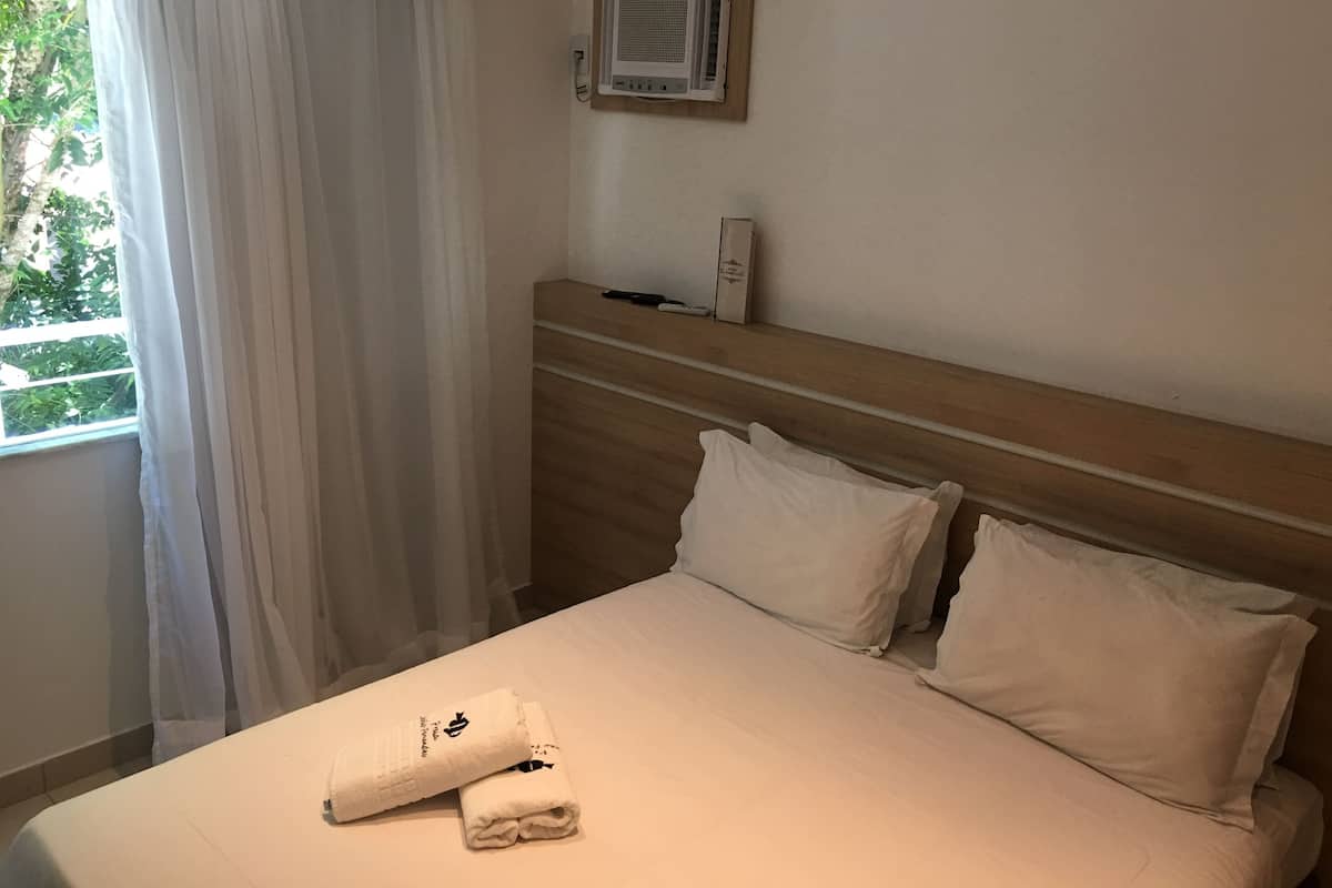 A hotel room with a bed, white curtains, a wooden headboard, and a wall-mounted air conditioner.