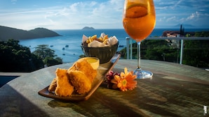 A table with a glass of iced drink, a plate of fried food, a bowl of sauce, and a flower, overlooking a scenic coastal view. - Pousada Praia João Fernandes (Búzios)