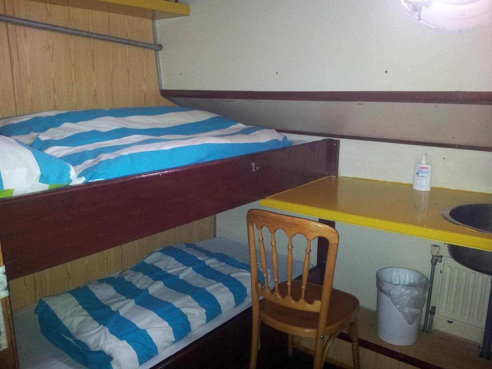 Una cama litera con ropa de cama a rayas azul y blanco, una silla de madera, una mesita con tapa amarilla, una botella blanca de desinfectante de manos y un bote de basura.