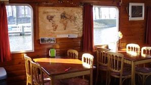 Un intérieur en bois, avec une table et des chaises en bois, une carte du monde accrochée au mur et une fenêtre donnant sur une étendue d’eau.