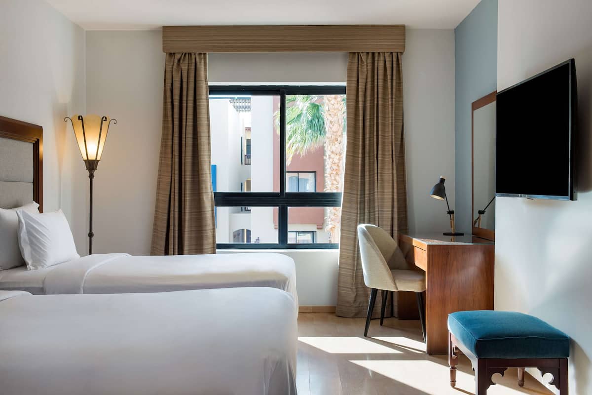 A hotel room with two beds, a desk, a chair, a TV, and a window with curtains.