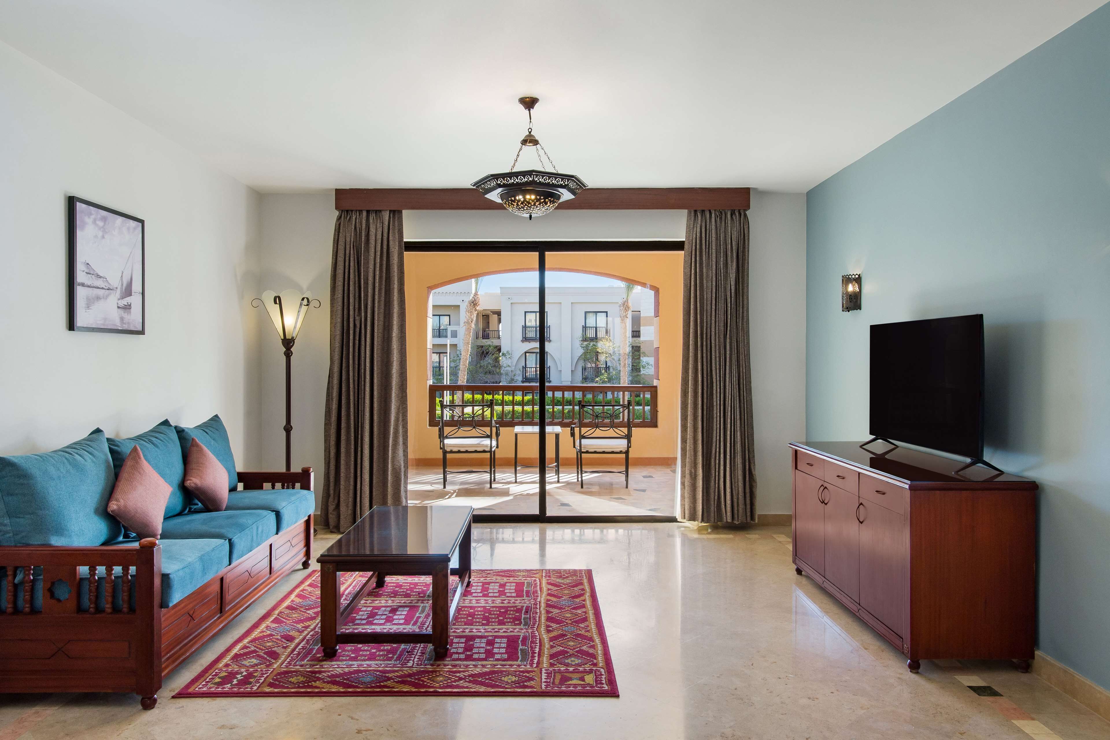 A living room with a sofa, a TV, a table, and a view of a balcony with outdoor furniture.