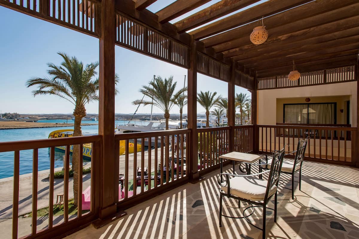 A covered outdoor area with a view of the marina, featuring a table and chairs, palm trees, and a wooden railing.