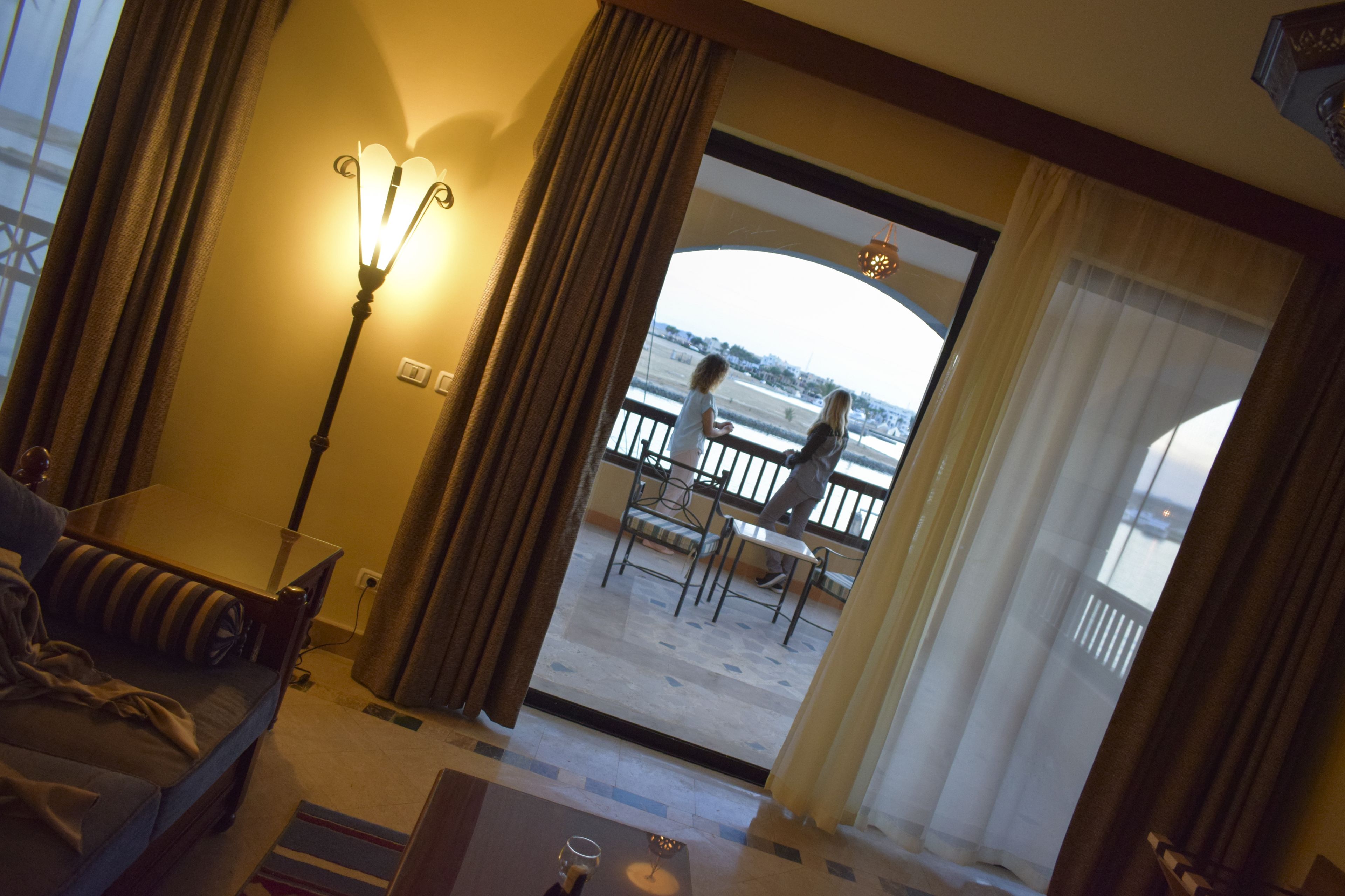 A hotel room with a sofa, a table, a lamp, and a view of a balcony with chairs.