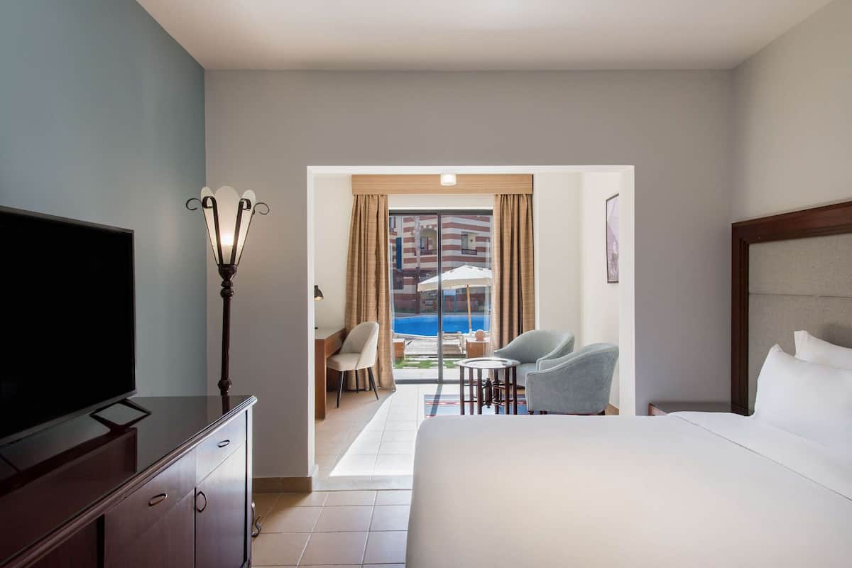 A hotel room with a bed, a TV, a desk, and a view of a pool and outdoor seating area.