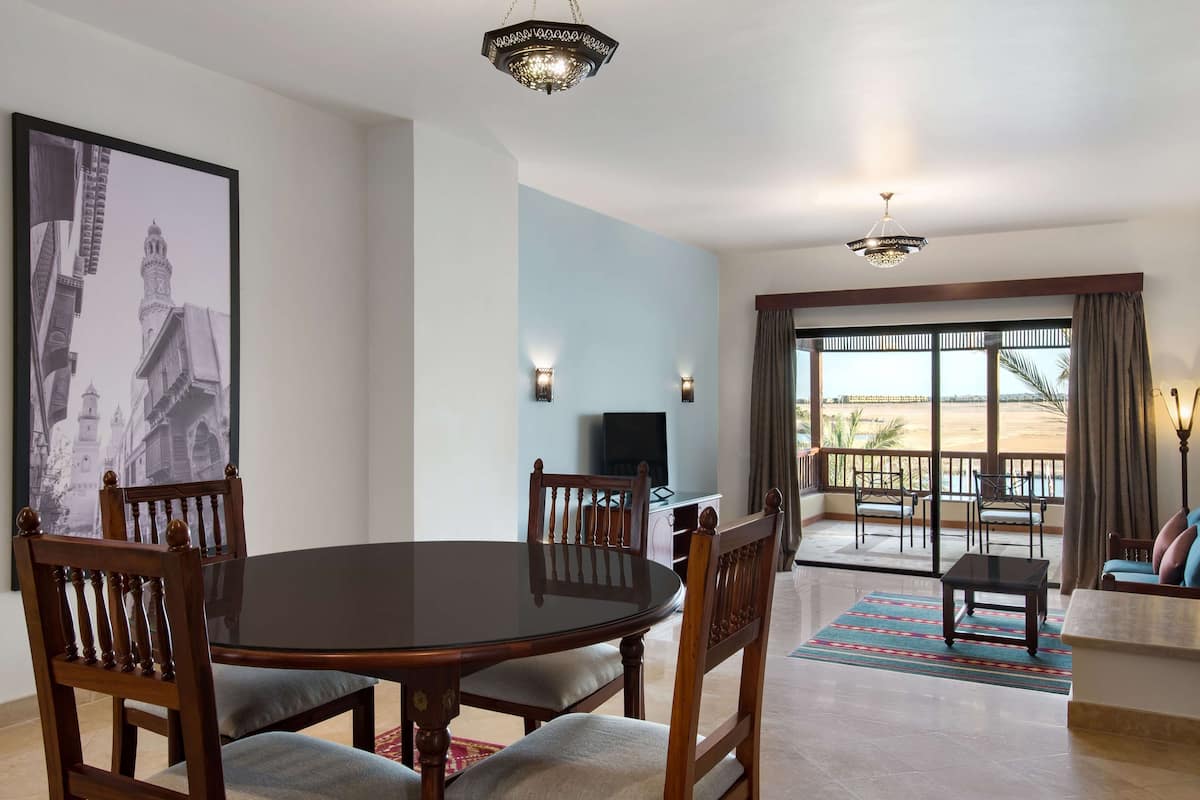 A dining area with a round table and chairs, a television, and a balcony with a view of the beach.