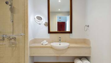 Un baño con ducha de azulejos color crema, lavamanos blanco con espejo arriba y dos toallas enrolladas sobre el mostrador.