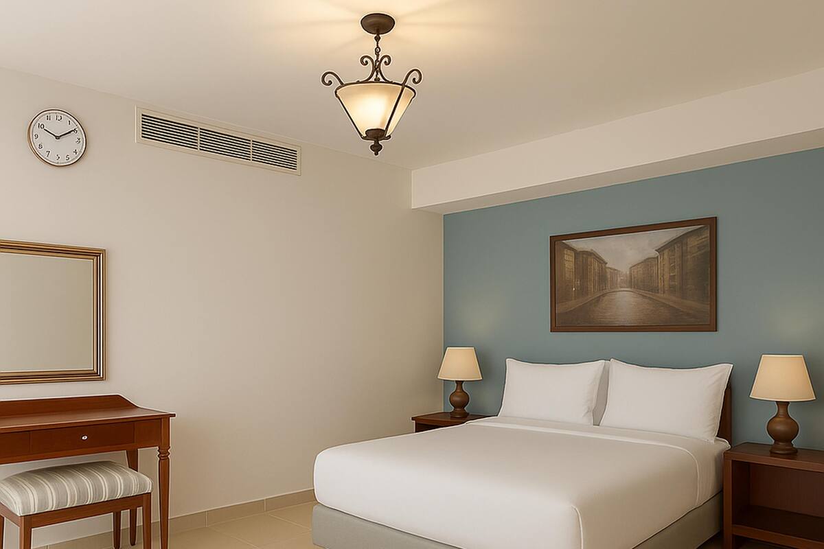 A hotel room with a bed, two bedside tables with lamps, a desk with a chair, a large mirror, a painting on the wall, and a ceiling light fixture.