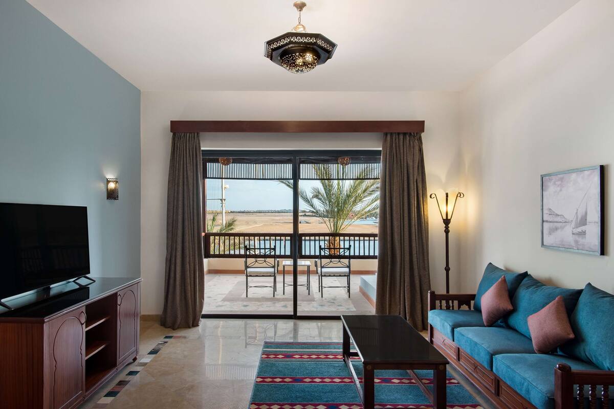 A living room with a TV, sofa, and a balcony with a view of the beach.