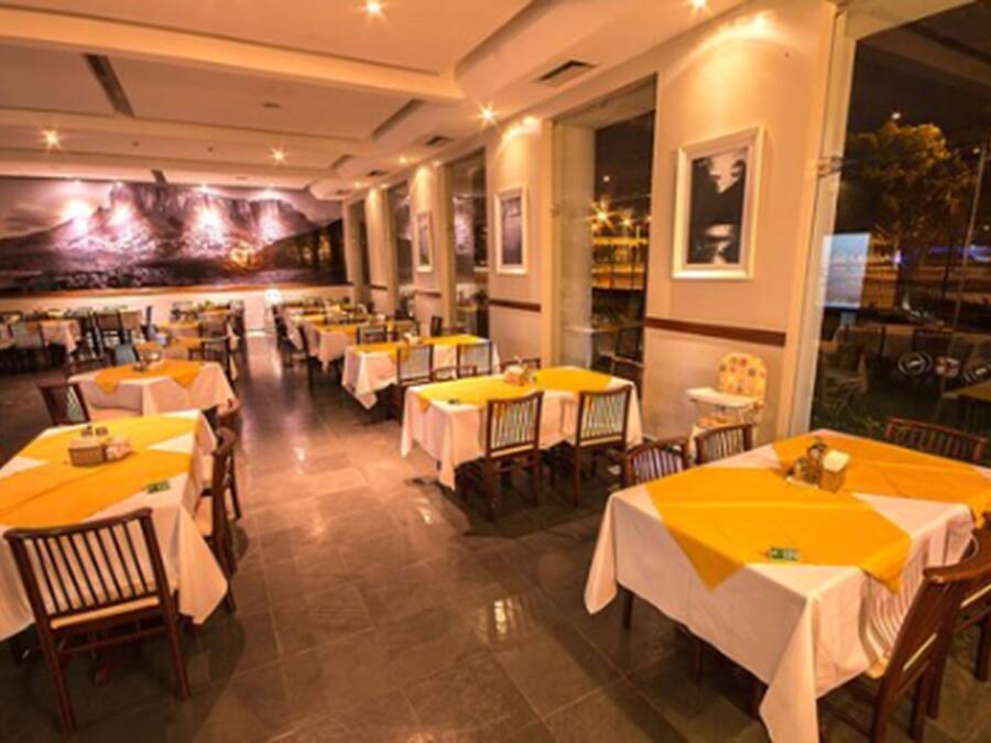A restaurant with multiple tables, each covered with a white tablecloth and a yellow runner. The room has a modern design with large windows and a mural on the wall.