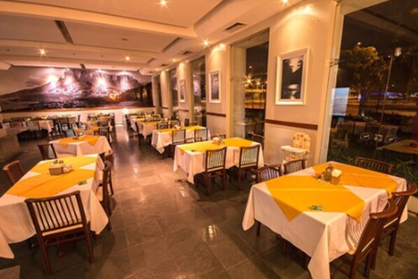 Un restaurante con varias mesas, cada una cubierta con mantel blanco y camino de mesa amarillo. El espacio tiene un diseño moderno, con ventanas amplias y un mural en la pared.