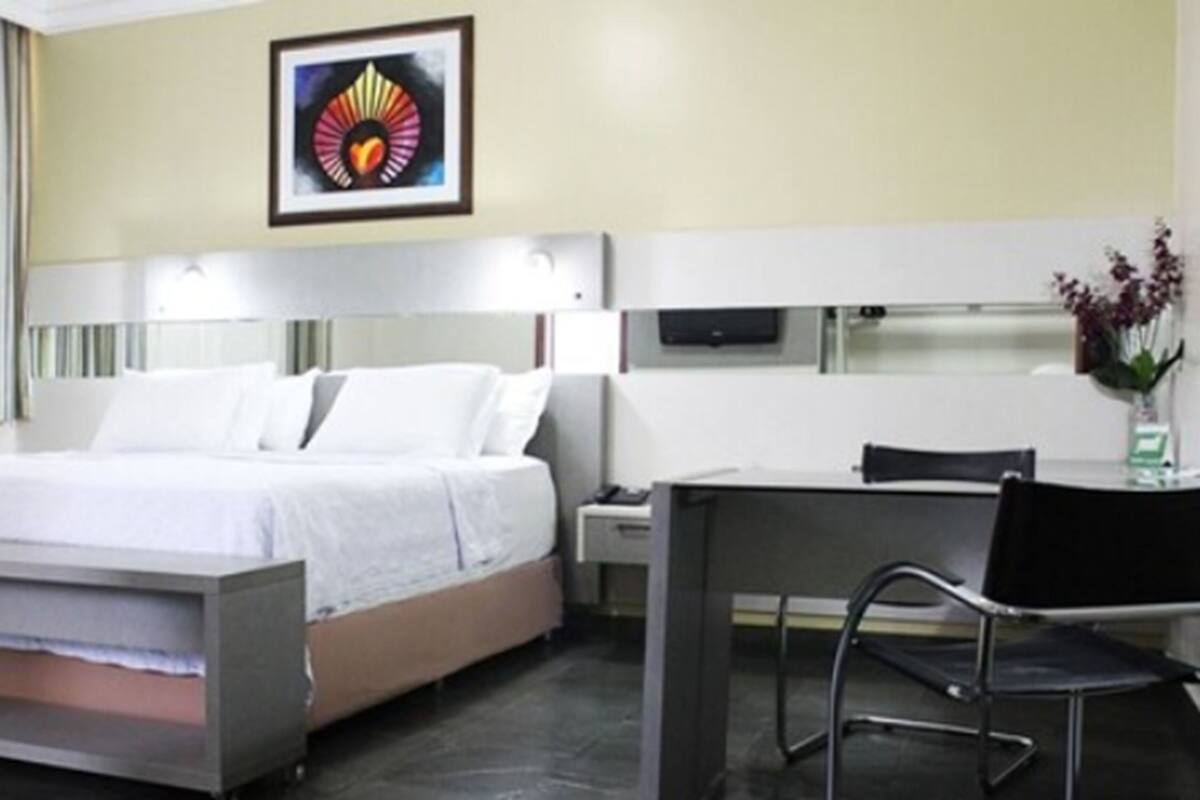 A hotel room with a bed, bedside table, chair, desk, and a painting on the wall.