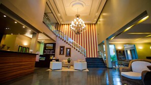 Een hotellobby met een receptiebalie, een zithoek, een trap en een kroonluchter.