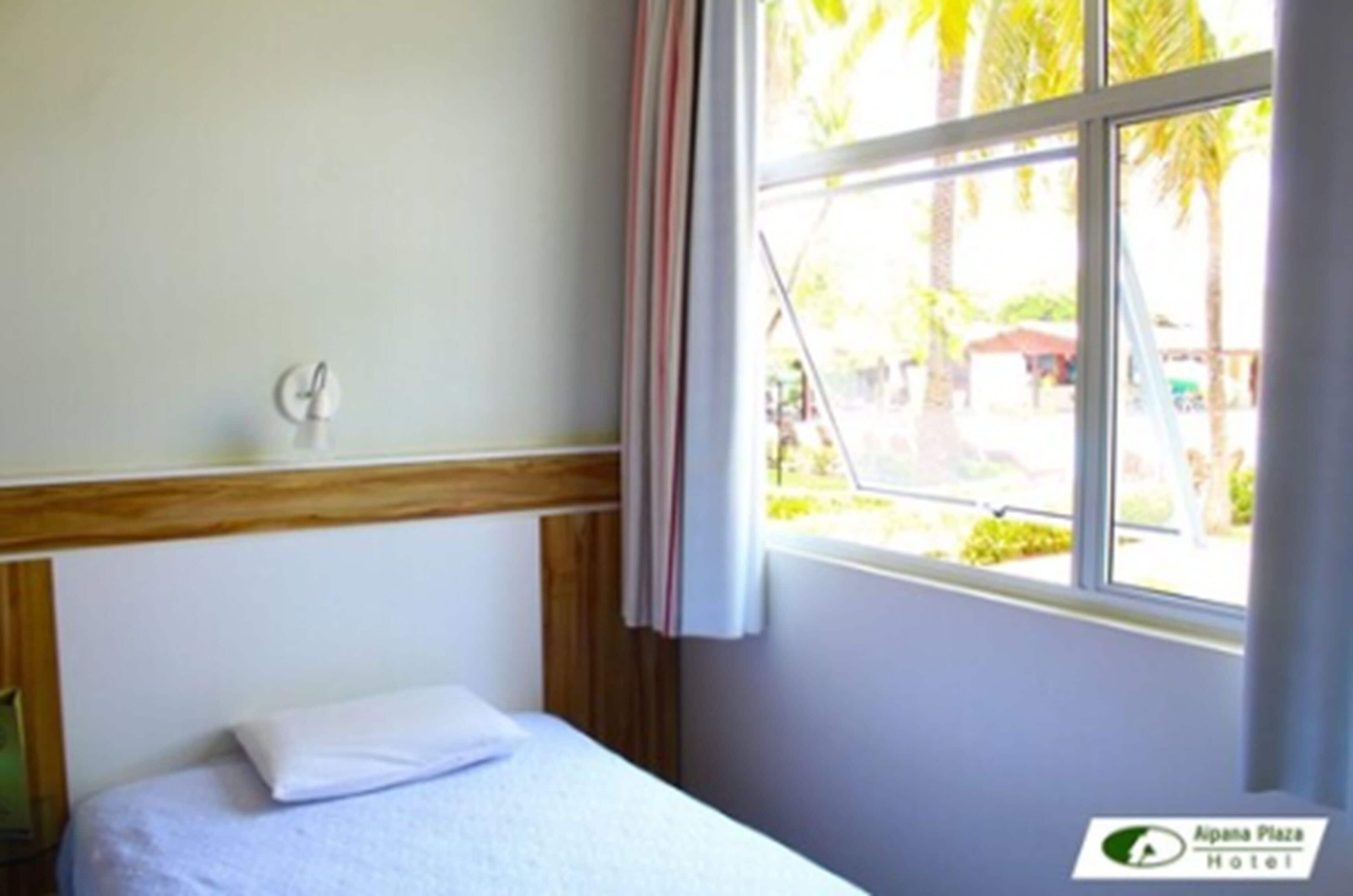 A hotel room with a bed, a window with curtains, and a view of palm trees outside.