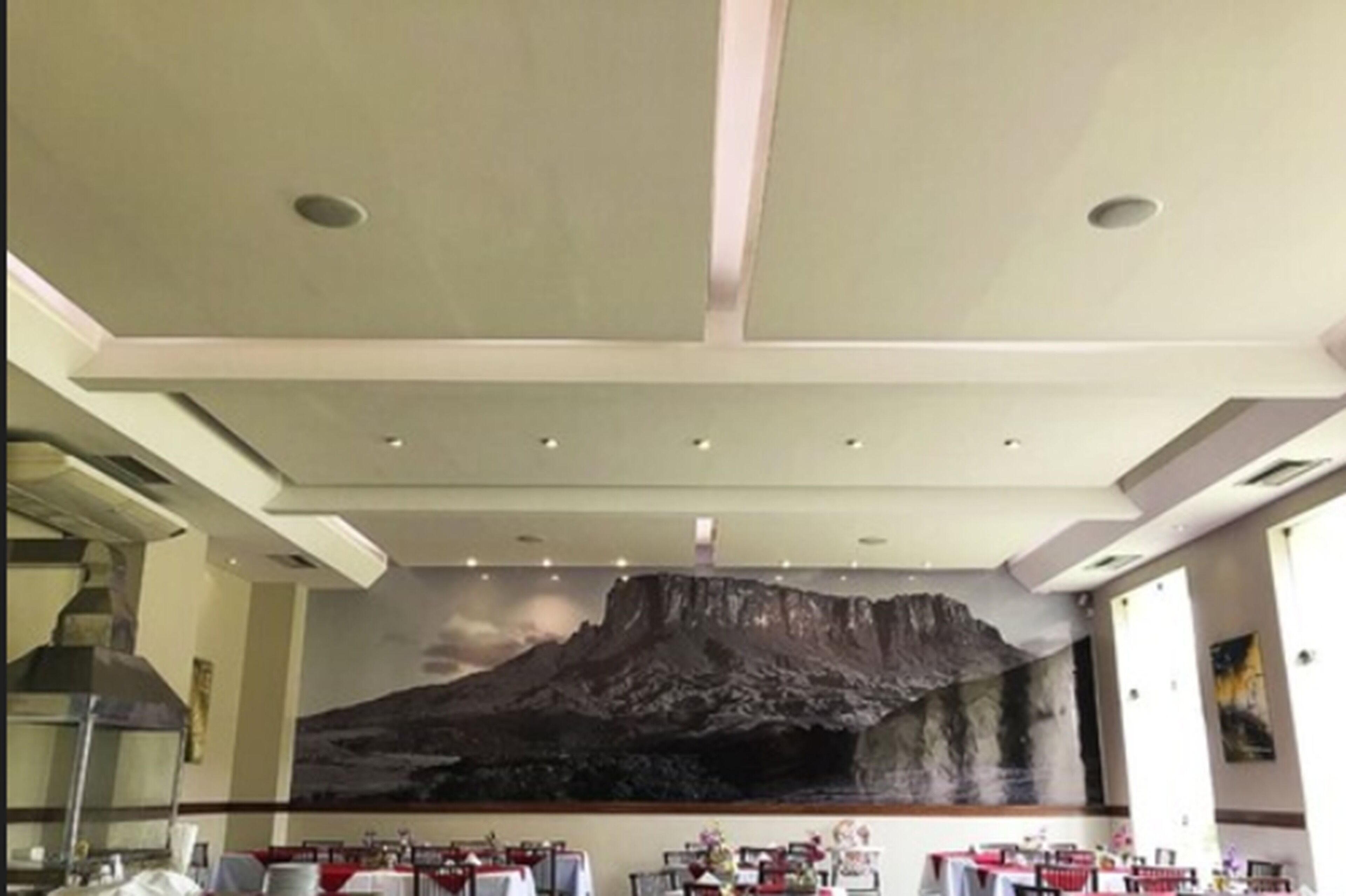 A restaurant with a high ceiling, recessed lighting, and a large mural of a mountain. Tables are set with white tablecloths and red chairs.