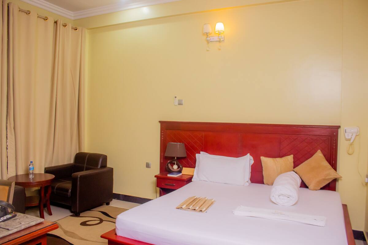 A hotel room with a bed, a chair, a table, a lamp, and a wall-mounted phone.