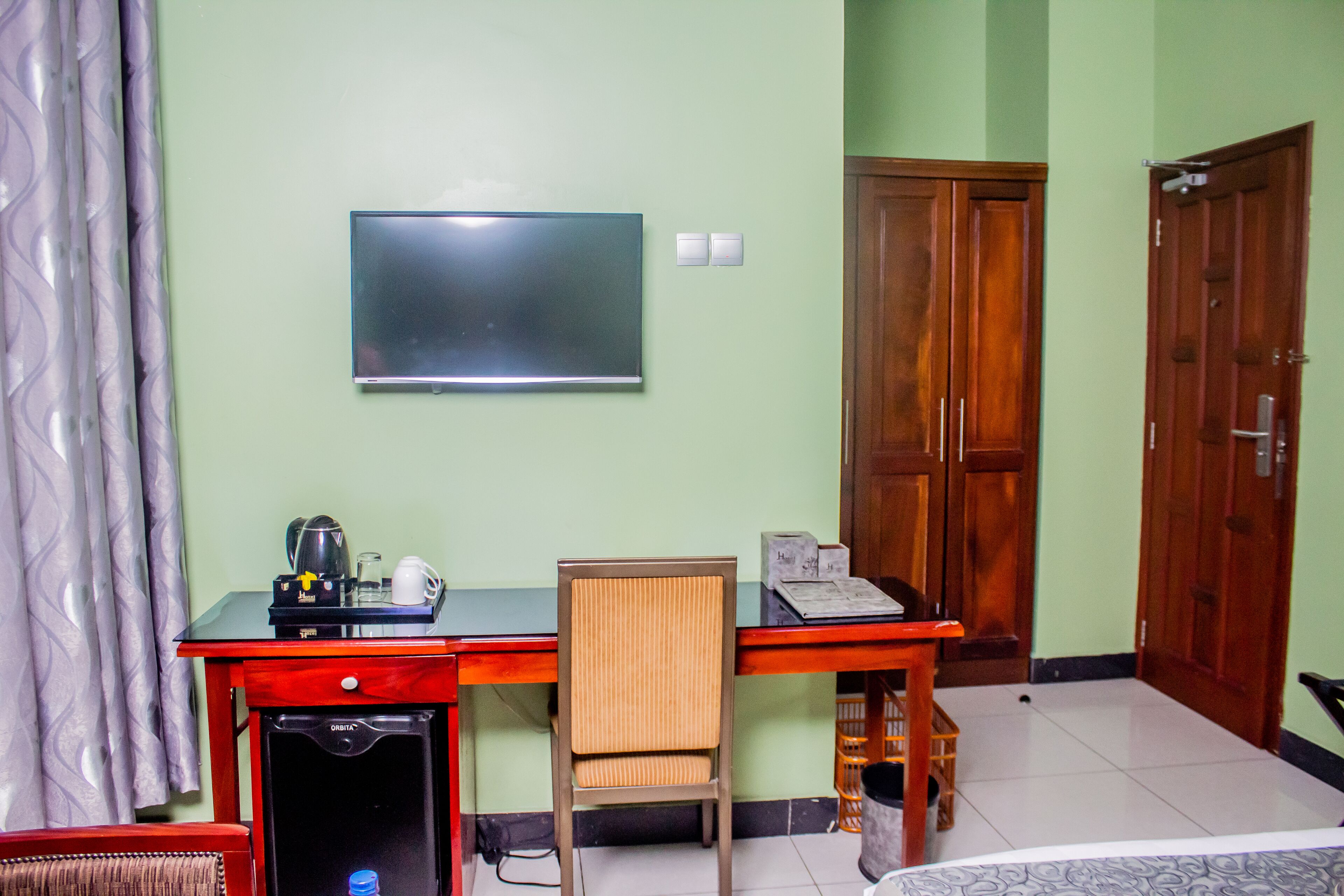 A room with a desk, chair, wardrobe, and a television on the wall.