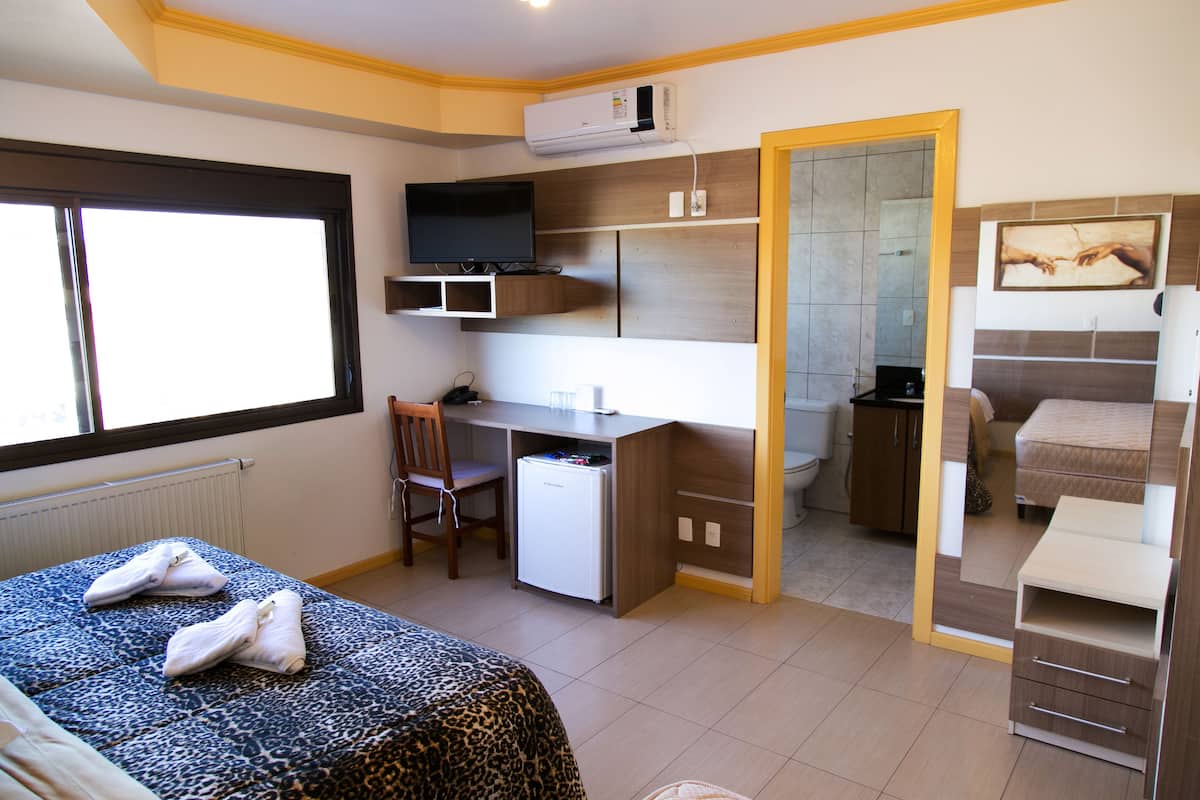 A hotel room with a bed, a desk, a chair, a small refrigerator, a television, and a bathroom visible through an open door.