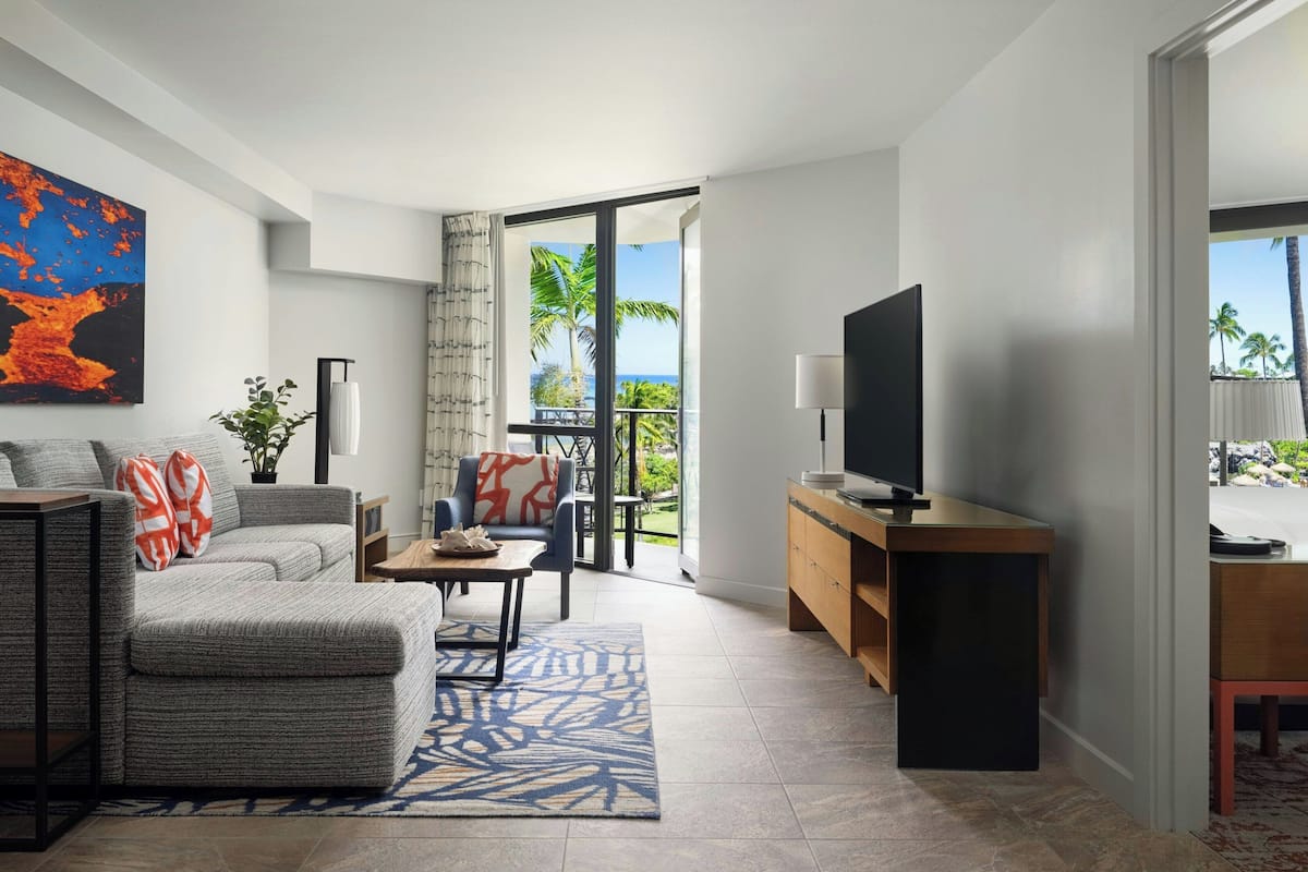 A modern living room with a grey sofa, a coffee table, and a television. There is a balcony with a view of palm trees and the ocean.