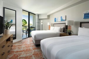 Habitación de hotel con dos camas, televisión, escritorio y balcón con vistas a palmeras.
