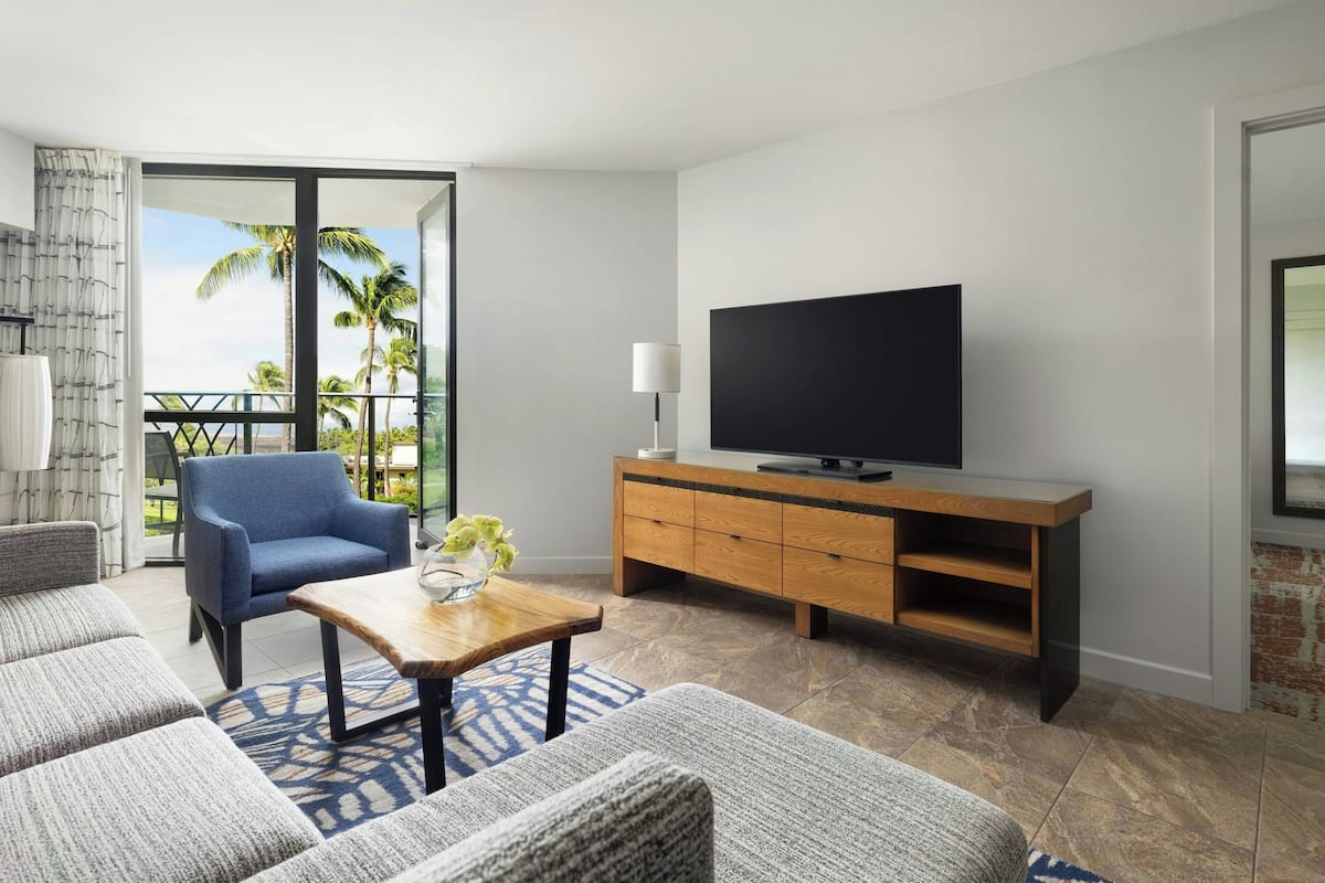 A modern living room with a sofa, a chair, a coffee table, a TV, and a balcony with a view of palm trees.