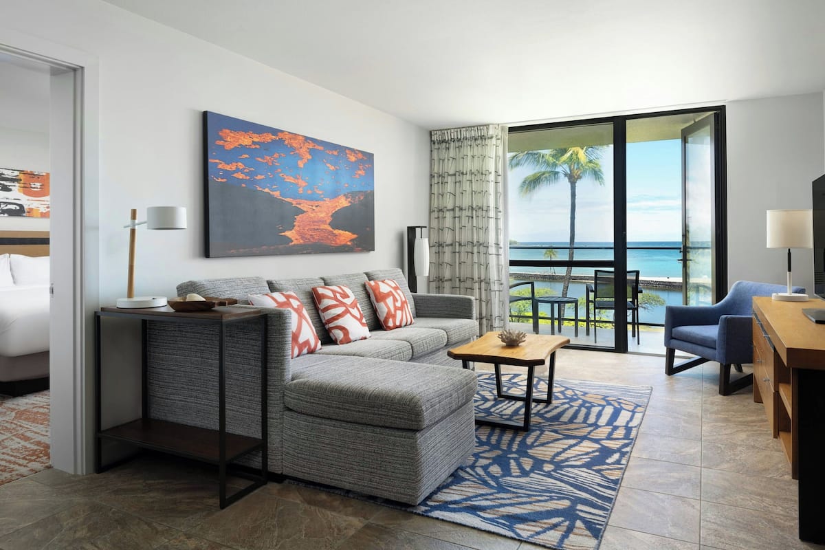 A modern living room with a grey sofa, a wooden coffee table, and a view of the ocean through large windows.
