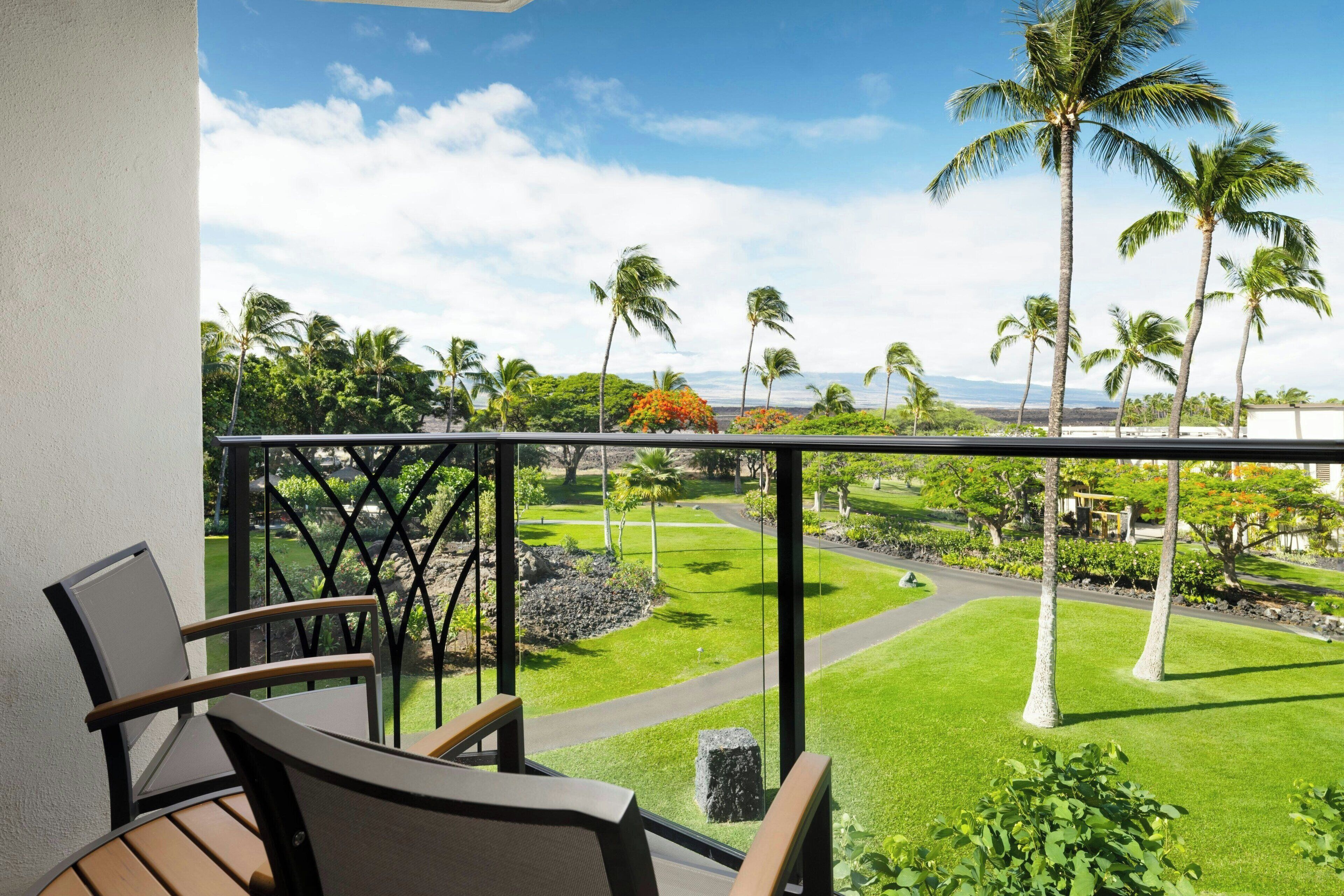 A balcony with a view of a well-maintained lawn, palm trees, and a distant landscape.