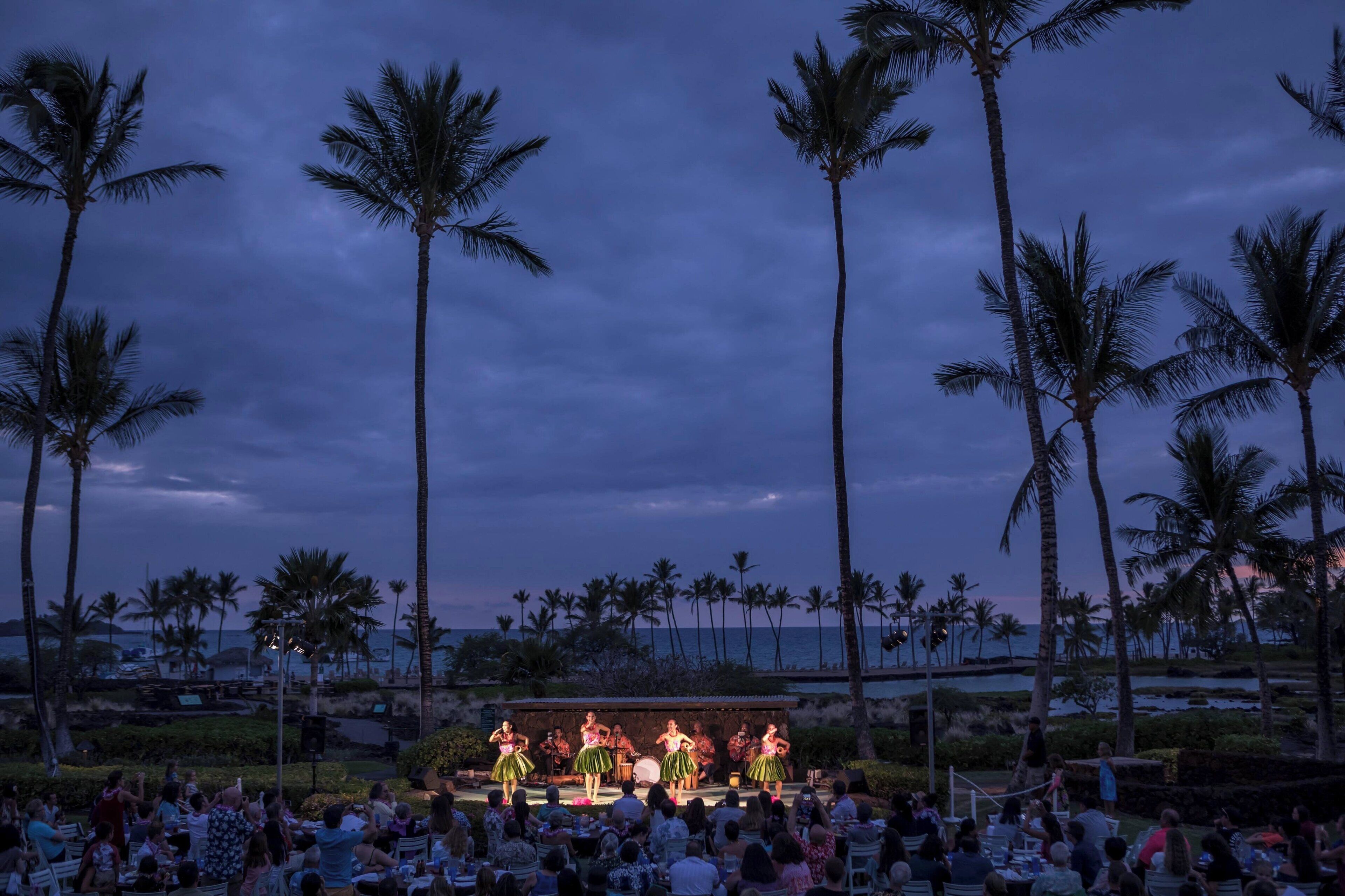 A stage with performers in traditional attire, surrounded by an audience, palm trees, and a view of the ocean.