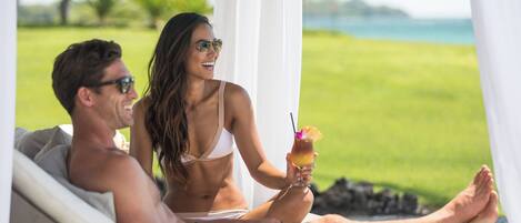 A couple relaxing on a sun lounger by a pool with a drink in hand, overlooking a grassy area and water.