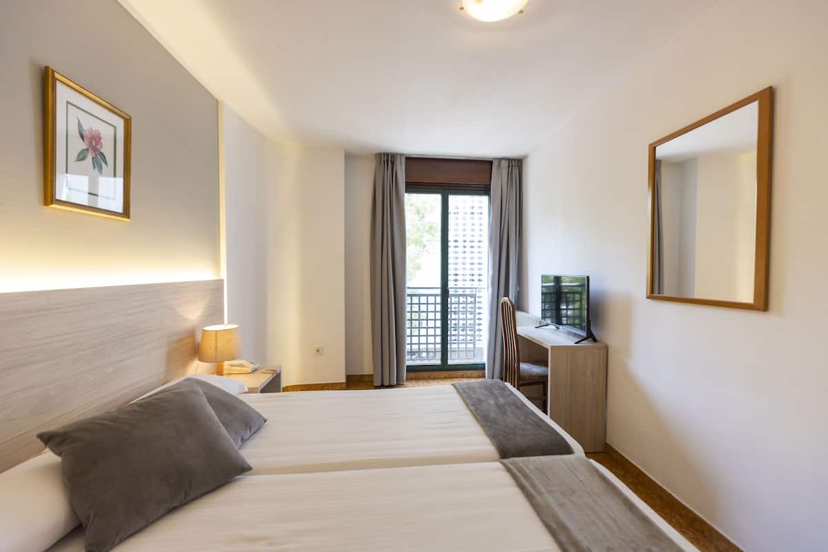 A hotel room with two beds, a desk, a chair, a TV, and a balcony with a view.
