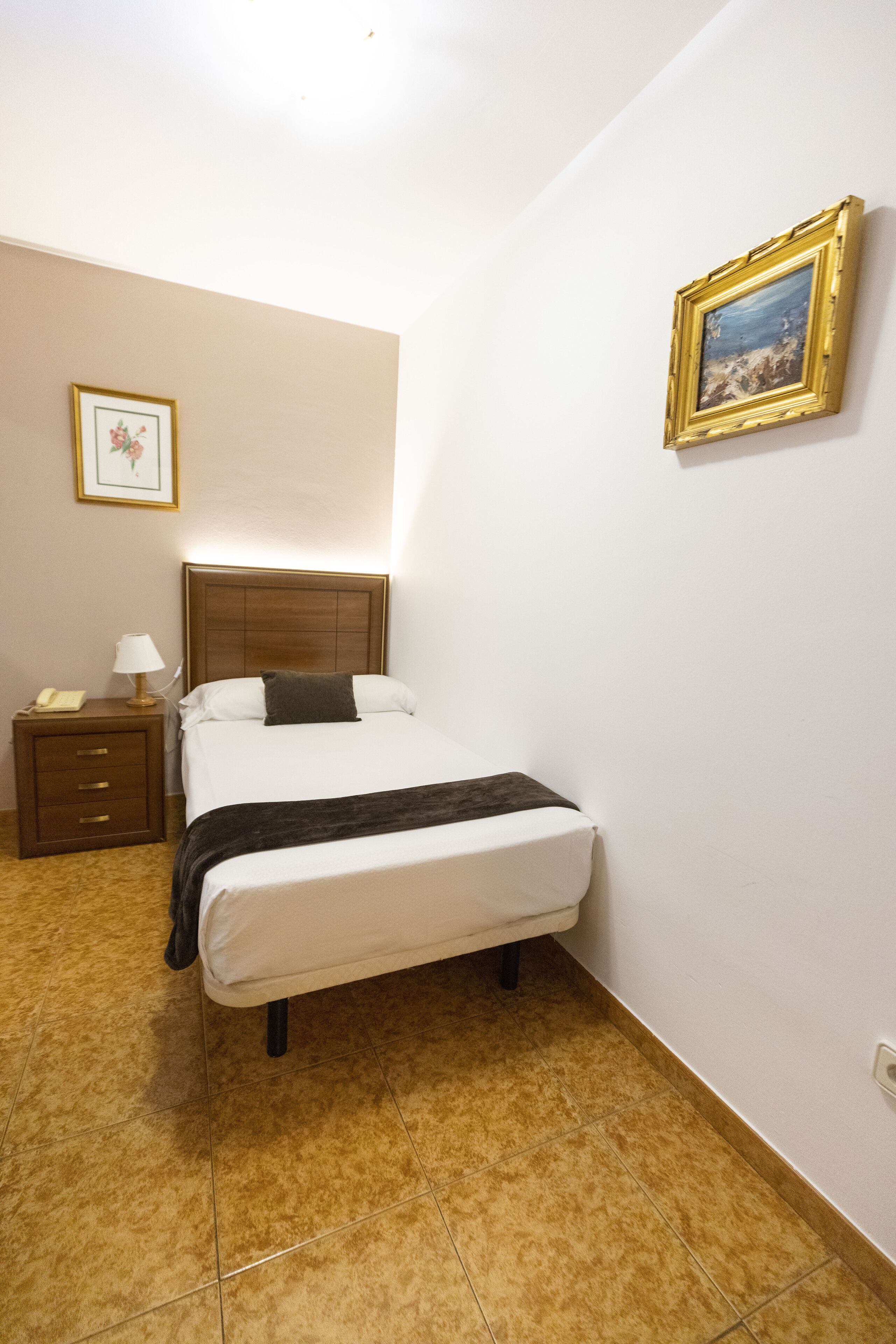 A hotel room with a bed, a nightstand, a lamp, a framed picture on the wall, and a tiled floor.