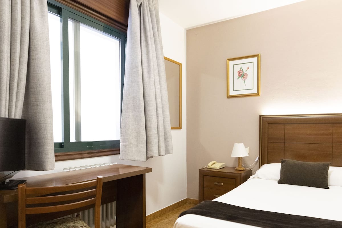 A hotel room with a bed, a desk, a chair, a nightstand, and a window with curtains.