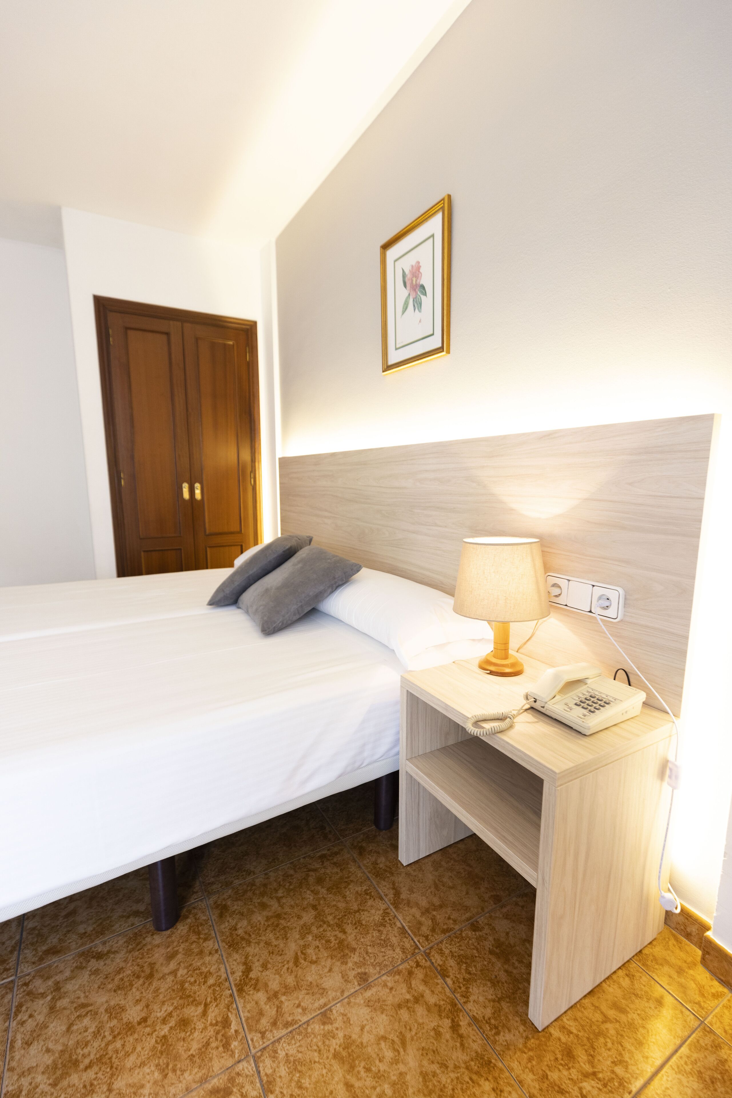 A hotel room with a bed, bedside table, lamp, phone, and a door.