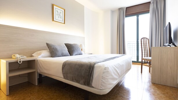 A hotel room with a bed, bedside table, a desk with a chair, a TV, and a window with curtains. - Hotel Madrid (Pontevedra)