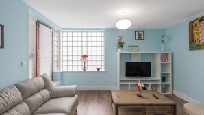 A living room with a beige sofa, a wooden coffee table, a TV on a white stand, and a window with blinds.