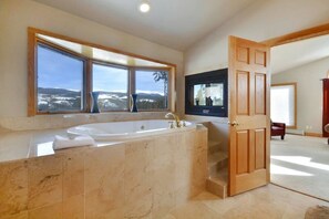 A bathroom with a large bathtub, a window with a view of snow-covered hills, and a wooden door. - Eagle Wind Lodge 5 Bedroom Holiday Home by Winter Park Lodging Company (Winter Park)