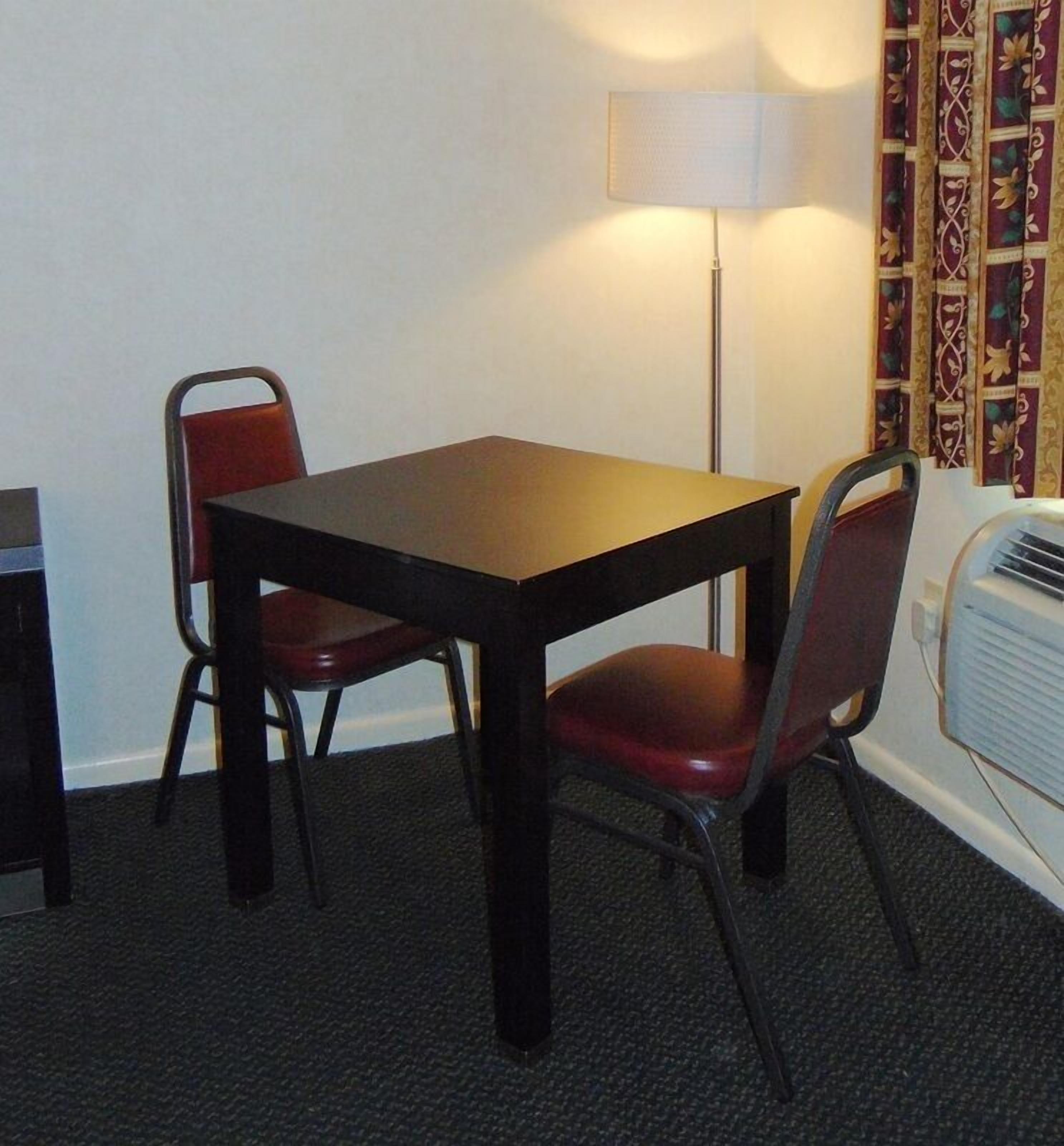 A small, dark wooden table with two red chairs, a floor lamp, and patterned curtains in a room with carpeted flooring.